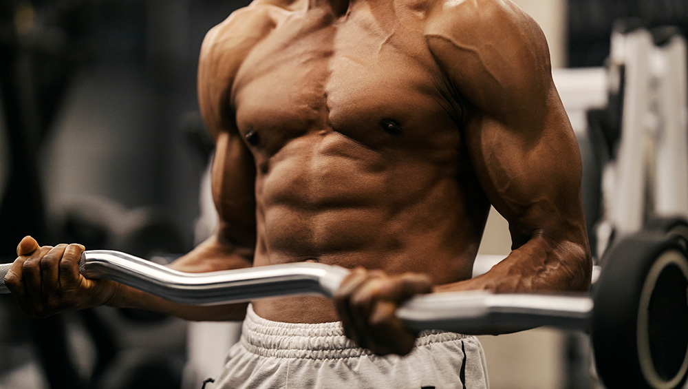 Shirtless muscular multicultural bodybuilder doing biceps curls at gym.