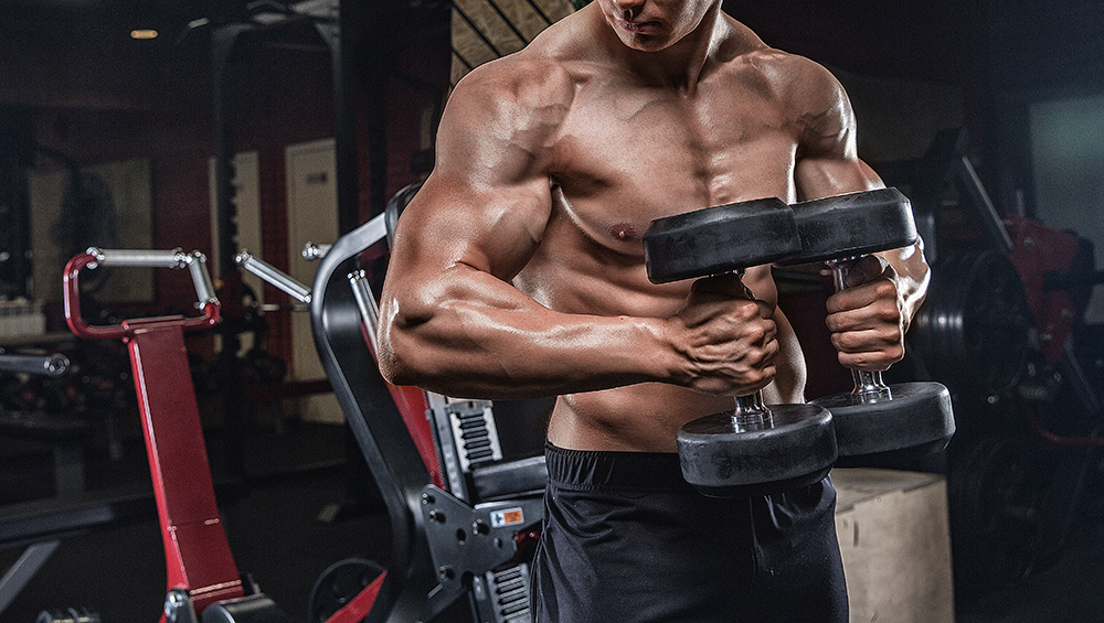 Muscular shirtless guy holding two dumbells in a dark gym